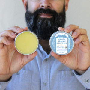 Man with a beard holding up a tin of Dr. Bronner's Unscented Magic Balm.