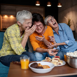 Three women friends sitting on a sofa with snacks laughing.