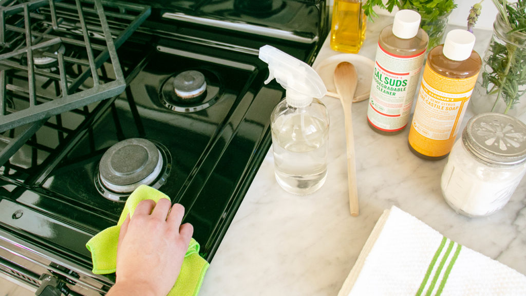 A person is cleaning a stovetop with Dr. Bronner's products and other natural cleaning products in the frame. how to clean an oven and stovetop