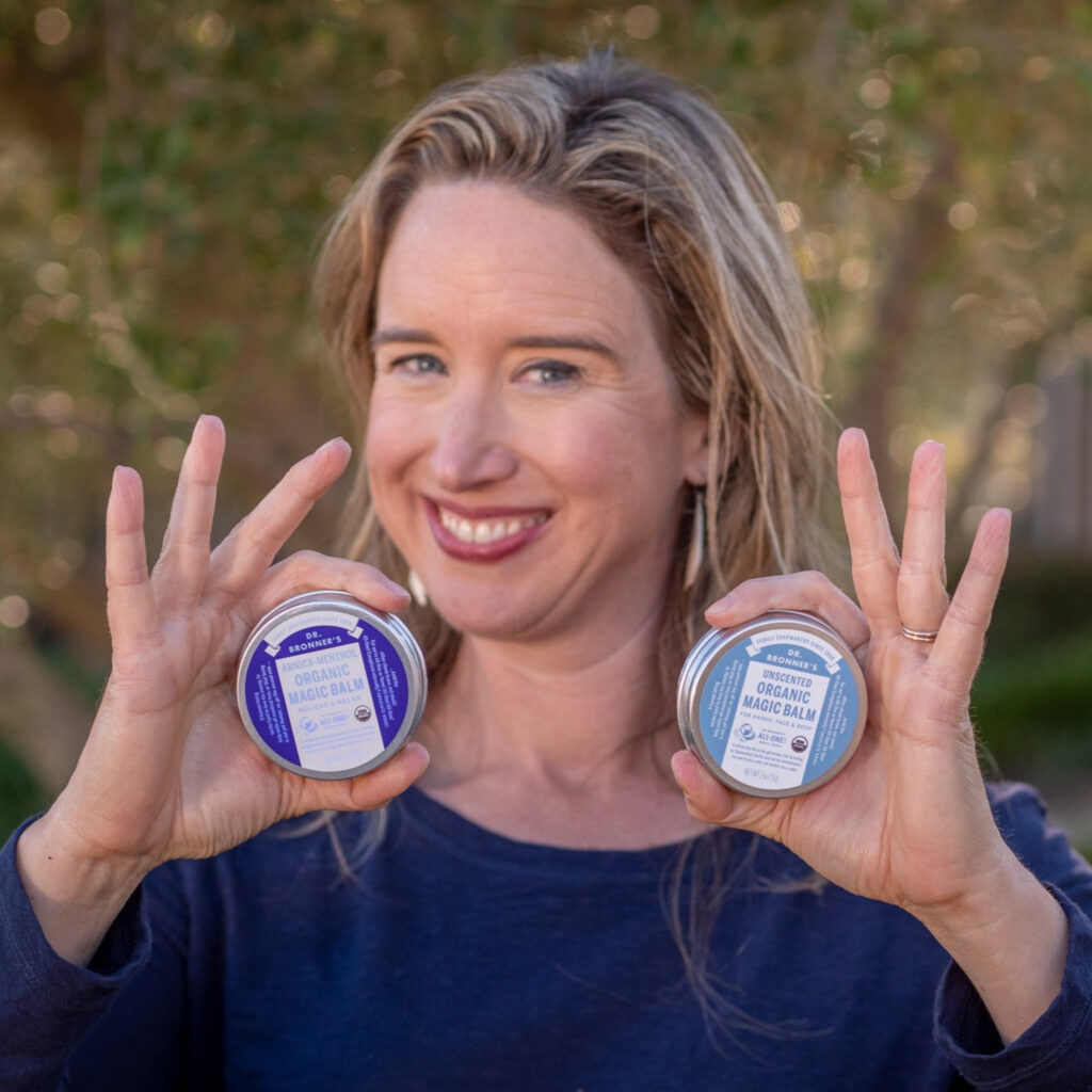 Lisa Bronner holding Dr. Bronner's Unscented Magic Balm and Arnica-Menthol Magic Balm - Usage Cheat Sheet