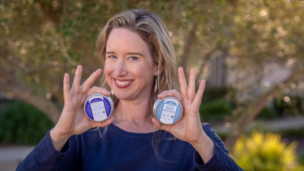 Lisa Bronner holding Dr. Bronner's Unscented Magic Balm and Arnica-Menthol Magic Balm - Usage Cheat Sheet