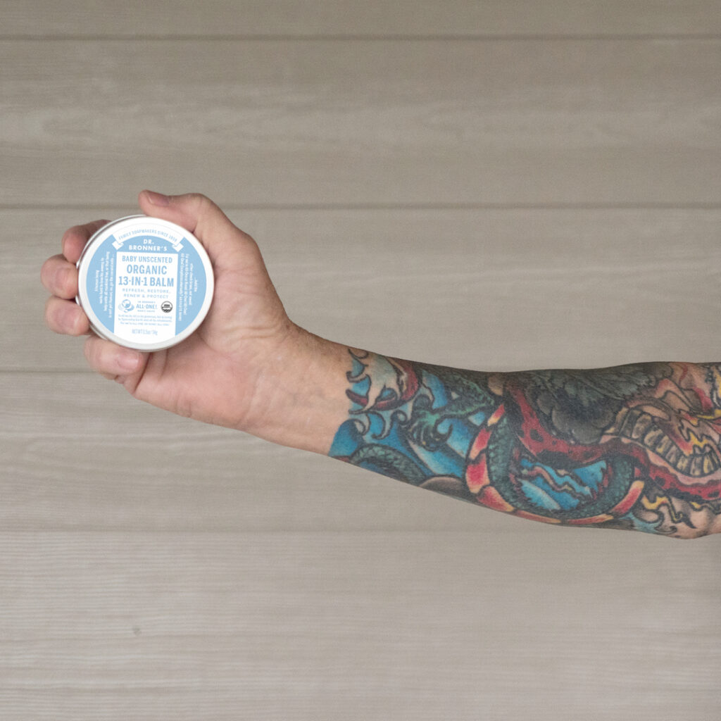 Person with tattoo sleeve holding Dr. Bronner's Unscented Magic Balm - tattoo care