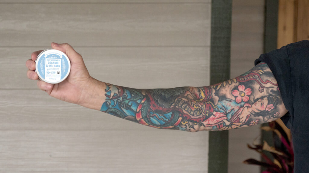 Person with tattoo sleeve holding Dr. Bronner's Unscented Magic Balm - tattoo care
