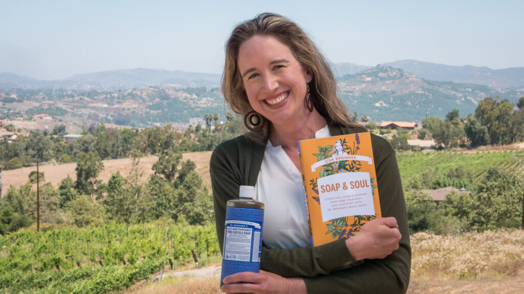 Lisa Bronner holding a copy of her book, Soap & Soul, and Dr. Bronner's Peppermint Castile Soap