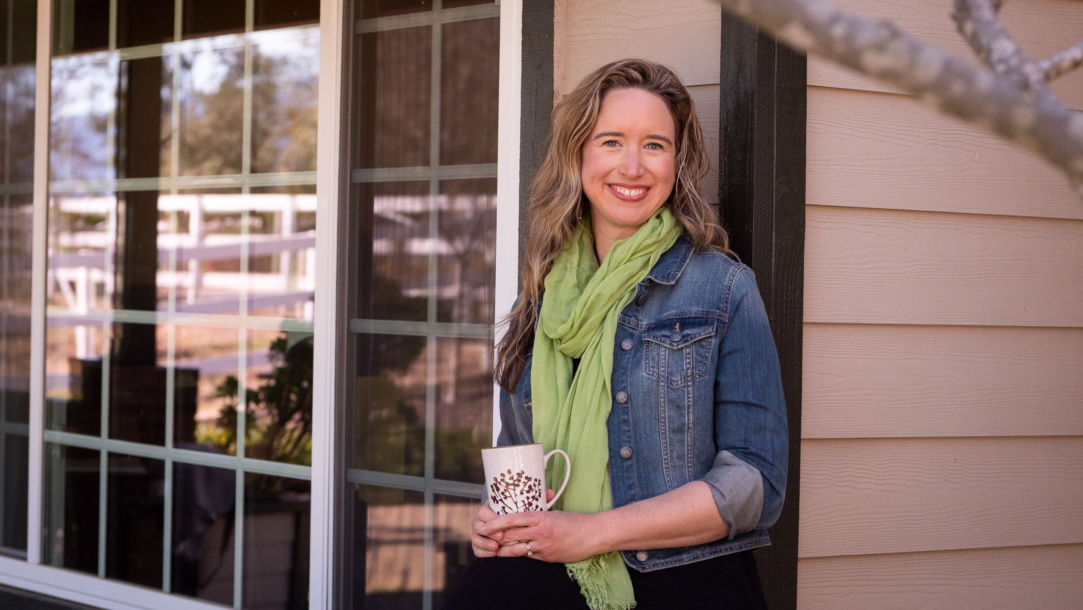 Lisa Bronner smiling, holding a coffee up in front of a house.