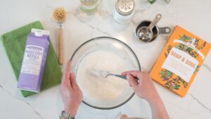 A bowl of GIY Soft Scrub surrounded by the ingredients to make it including Dr. Bronner's Castile Magic Soap.