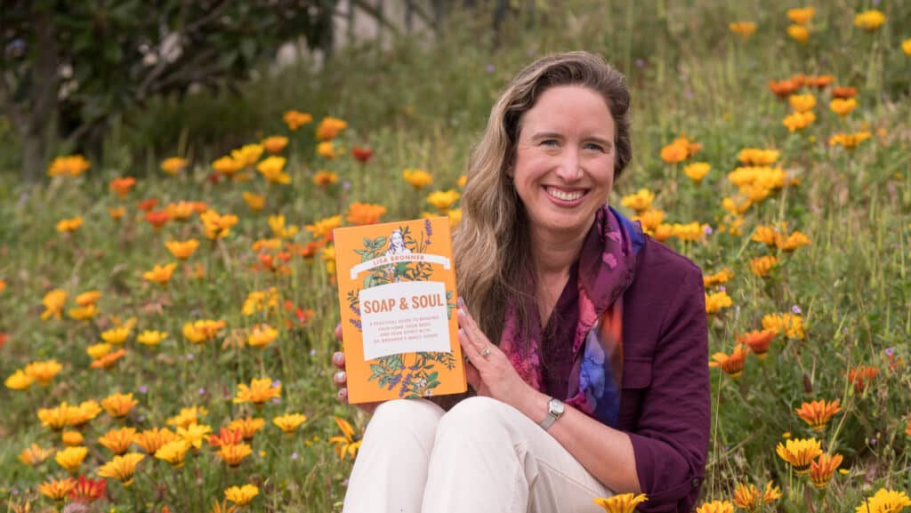 Lisa sitting in flower field, smiling, holding up a copy of her book Soap & Soul.